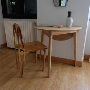 Puede incluir: Una mesa de madera con un dise&ntilde;o redondeado de hoja ca&iacute;da y una silla de madera. La mesa tiene un acabado de madera clara y la silla tiene un acabado de madera oscura. La mesa est&aacute; puesta con un peque&ntilde;o cuenco rosa y un jarr&oacute;n con un dise&ntilde;o marr&oacute;n y blanco.