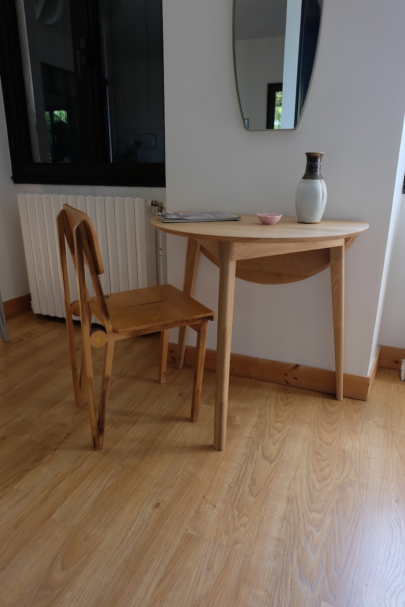 Puede incluir: Una mesa de madera con un dise&ntilde;o redondeado de hoja ca&iacute;da y una silla de madera. La mesa tiene un acabado de madera clara y la silla tiene un acabado de madera oscura. La mesa est&aacute; puesta con un peque&ntilde;o cuenco rosa y un jarr&oacute;n con un dise&ntilde;o marr&oacute;n y blanco.