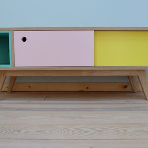 Op de afbeelding: Een houten kast met drie lades, één turquoise, één roze en één geel. De kast heeft een mid-century modern stijl met taps toelopende poten.