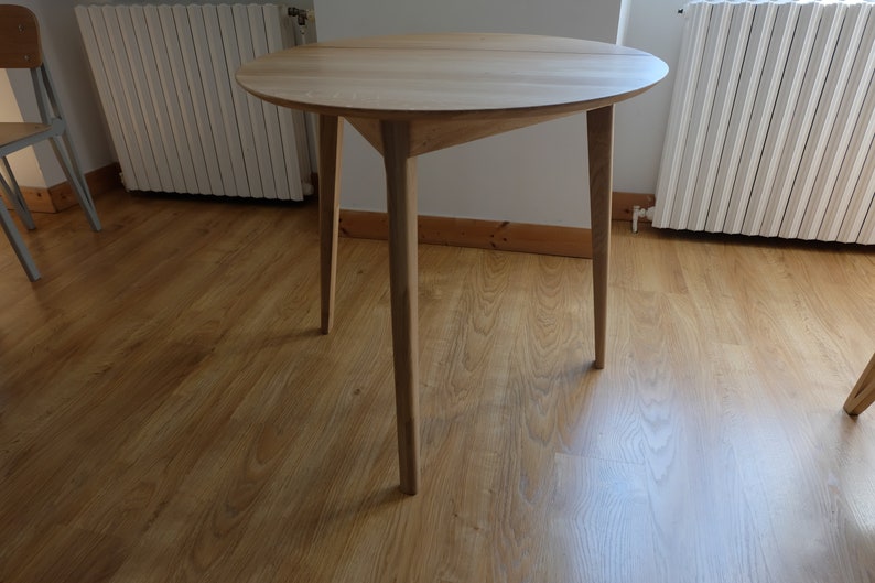 Puede incluir: Una mesa redonda de madera con un acabado de madera clara y tres patas. La mesa est&aacute; sobre un suelo de madera dura.