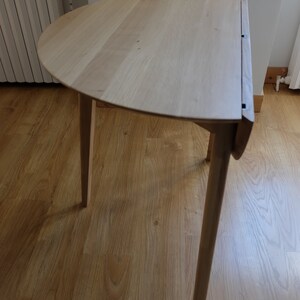 Puede incluir: Una mesa plegable de madera clara con una tapa semicircular y tres patas. La mesa est&aacute; sobre un suelo de madera.