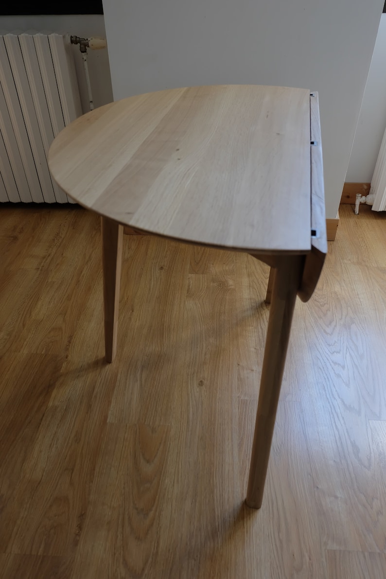 Puede incluir: Una mesa plegable de madera clara con una tapa semicircular y tres patas. La mesa est&aacute; sobre un suelo de madera.