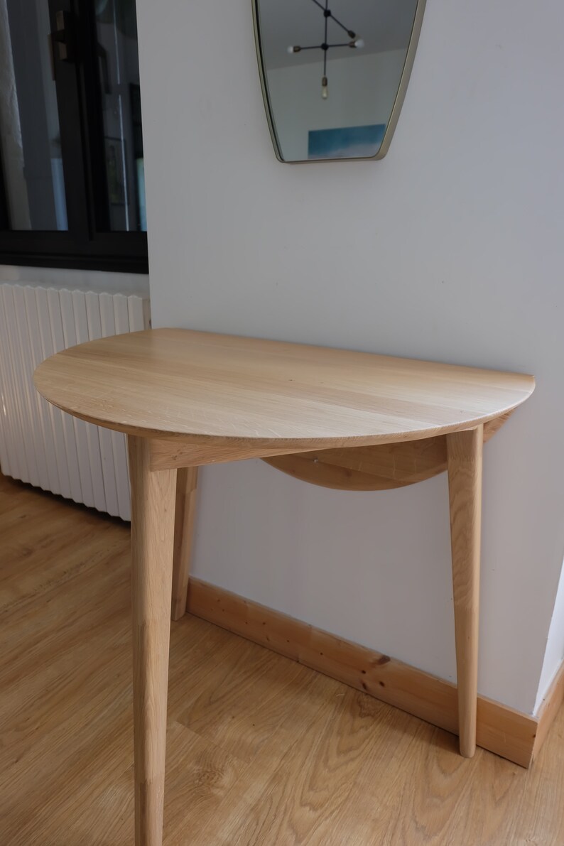 Puede incluir: Una mesa de madera clara de media luna con una tapa curva y tres patas. La mesa est&aacute; hecha de madera maciza y tiene un acabado natural.