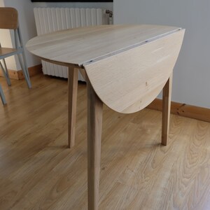 Puede incluir: Una mesa plegable de madera clara con una tapa redonda y cuatro patas. La mesa est&aacute; cerrada y la tapa est&aacute; doblada por la mitad.