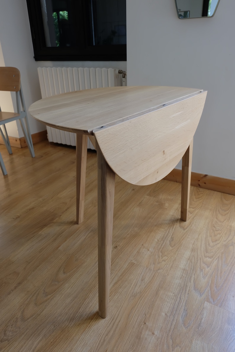Puede incluir: Una mesa plegable de madera clara con una tapa redonda y cuatro patas. La mesa est&aacute; cerrada y la tapa est&aacute; doblada por la mitad.