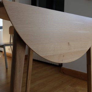 Puede incluir: Una mesa plegable de madera clara con una tapa redonda semicircular y cuatro patas. La mesa est&aacute; cerrada y la tapa est&aacute; doblada por la mitad.