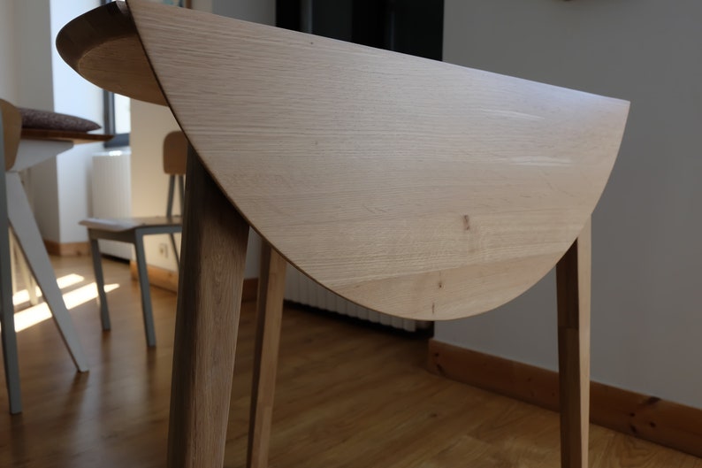 Puede incluir: Una mesa plegable de madera clara con una tapa redonda semicircular y cuatro patas. La mesa est&aacute; cerrada y la tapa est&aacute; doblada por la mitad.