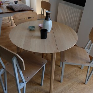 Puede incluir: Una mesa redonda de madera clara con una tapa de madera clara y cuatro patas. La mesa est&aacute; puesta con dos cuencos peque&ntilde;os y una taza negra. Hay tres sillas alrededor de la mesa, dos con asientos de madera clara y patas de metal gris, y una con un asiento de madera clara y patas de madera oscura.