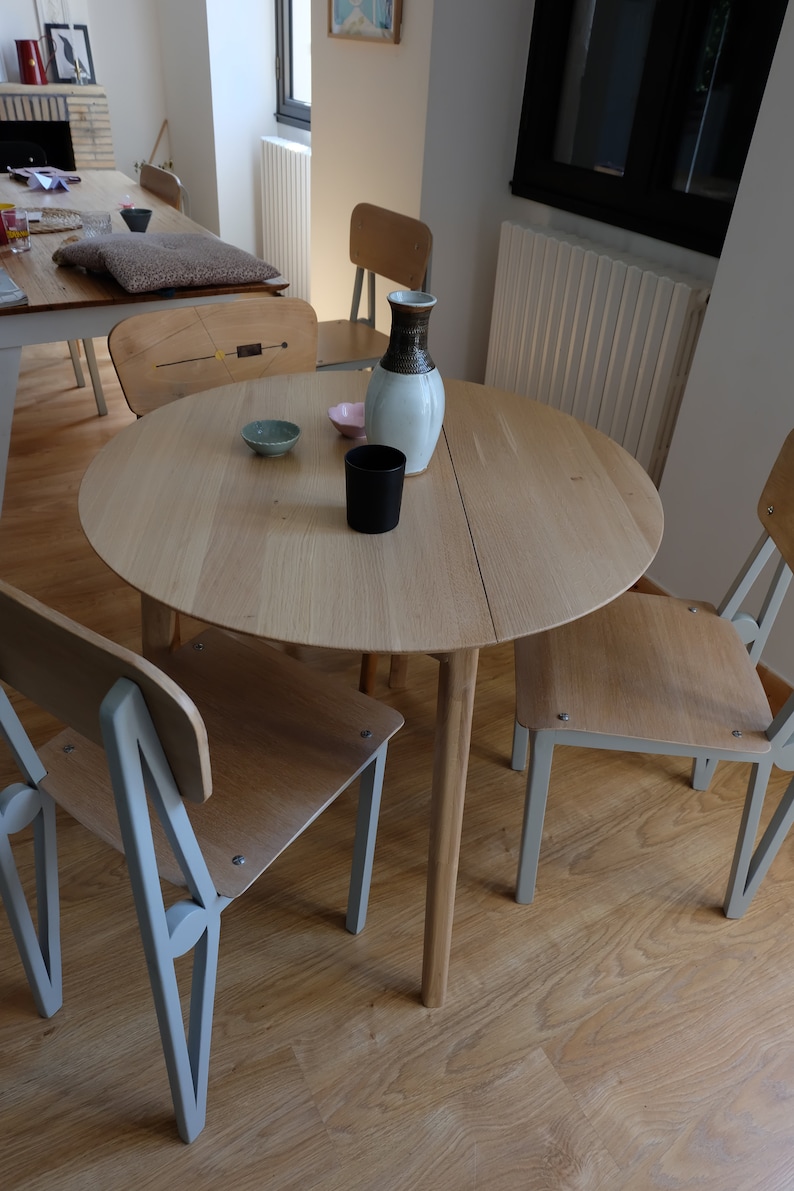 Puede incluir: Una mesa redonda de madera clara con una tapa de madera clara y cuatro patas. La mesa est&aacute; puesta con dos cuencos peque&ntilde;os y una taza negra. Hay tres sillas alrededor de la mesa, dos con asientos de madera clara y patas de metal gris, y una con un asiento de madera clara y patas de madera oscura.