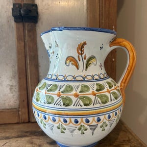 May include: A white ceramic pitcher featuring a hand-painted design with green leaf motifs, blue scalloped borders, and yellow accents. The vessel has a wide, rounded body, a flared rim with a pouring spout, and a solid orange-brown curved handle.