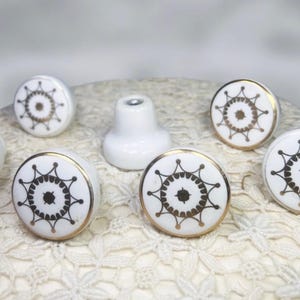May include: White ceramic cabinet knobs with a gold rim and a black and gold starburst design. The knobs are round and have a decorative pattern. A white ceramic cabinet pull is also present.