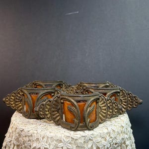 May include: A set of four circular napkin rings featuring an aged bronze-toned metal frame with intricate fan-shaped side detailing and a central amber-colored resin insert. Each piece displays a curved, ornate metal structure with leaf-like accents.
