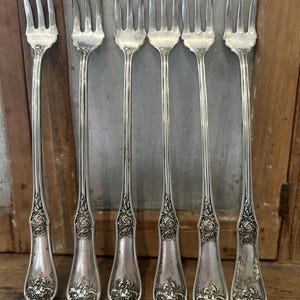 May include: A set of six silver-toned metal cocktail forks with long, slender handles. Each fork features a three-tined head and a decorative embossed floral pattern at the base of the handle, showcasing intricate leaf and petal detailing.