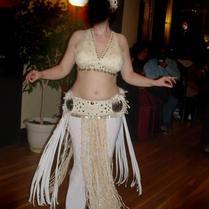 May include: A woman wearing a white belly dancing costume with a gold and white beaded belt and a white fringe skirt. She is wearing a white headpiece and has her hair pulled back.