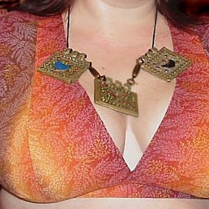 May include: A woman wearing a red, orange, and yellow patterned top with a gold necklace featuring three decorative pendants.
