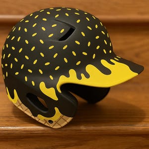 May include: Black baseball helmet with a yellow ice cream cone design. The helmet has a black base with yellow sprinkles and a yellow ice cream drip design on the brim. The ear flaps have a waffle cone design.