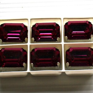 May include: Six dark purple rectangular rhinestones in a clear plastic packaging. The rhinestones are arranged in two rows of three.