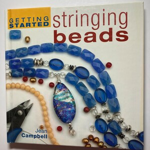 Może przedstawiać: Książka zatytułowana "Getting Started Stringing Beads" autorstwa Jean Campbell. Okładka przedstawia różne koraliki w kolorze niebieskim, białym, złotym i czerwonym. Koraliki są ułożone w okrągły wzór wokół dużej niebiesko-białej koraliki szklanej.