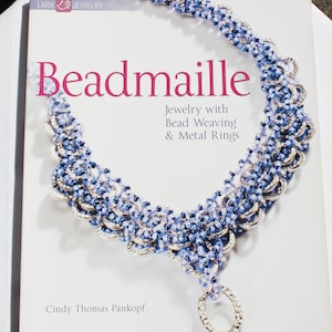 Puede incluir: Un libro titulado "Beadmaille: Joyería con tejido de cuentas y anillos de metal" de Cindy Thomas Pankopf, que presenta un primer plano de un collar de cuentas plateadas y azules con un colgante de anillo plateado.