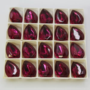 May include: A sheet of 20 clear, faceted, teardrop-shaped rhinestones in a deep magenta color. The rhinestones are arranged in a grid pattern in a white plastic tray.