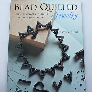 May include: A book titled "Bead Quilled Jewelry: New Beadwork Designs with Square Stitch" by Kathy King. The cover features a black beaded necklace with a zig-zag pattern.