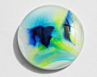 May include: A round, clear glass cabochon with a swirling design of blue, green, and yellow colors. The design includes a dark blue shape and glitter accents. The cabochon is set against a white background.