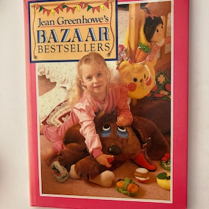 Könnte beinhalten: Ein rosa Buch mit dem Titel "Jean Greenhowe's Bazaar Bestsellers" mit einem Foto eines jungen Mädchens, das mit einem Stoffhund und anderen Spielzeugen spielt, auf dem Cover.