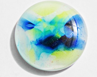 May include: A round, glass decorative object with a colorful abstract design. The design features swirls of blue, green, and yellow hues, with a touch of glitter. The object is set against a white background, creating a striking contrast.