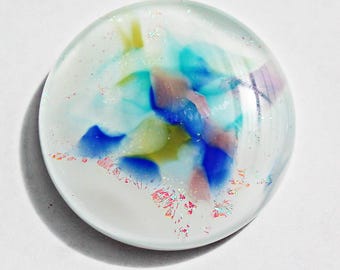 May include: A clear, round glass paperweight with colorful abstract art. The design features blue, teal, pink, and yellow shapes within the glass, with iridescent glitter accents. The paperweight is set against a white background.
