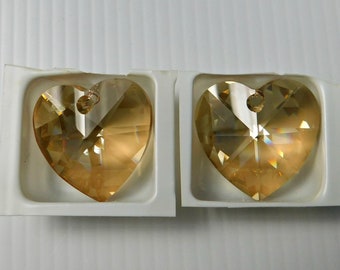 May include: Two gold heart-shaped crystal pendants with a faceted design. The pendants are displayed in white cardboard packaging.