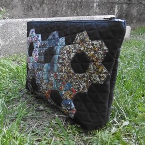 Pu&ograve; includere: Pochette in tessuto trapuntato nero con chiusura a cerniera. La pochette presenta un design patchwork di forme esagonali con vari motivi floreali. La borsa &egrave; appoggiata sull'erba verde.