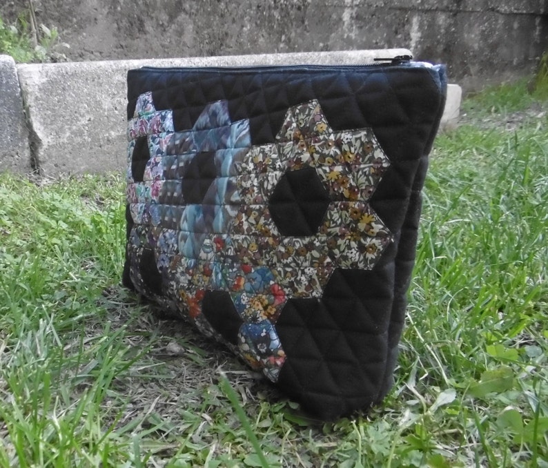 Pu&ograve; includere: Pochette in tessuto trapuntato nero con chiusura a cerniera. La pochette presenta un design patchwork di forme esagonali con vari motivi floreali. La borsa &egrave; appoggiata sull'erba verde.