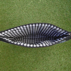 Pu&ograve; includere: Pochette nera con cerniera e fodera a quadretti bianchi e neri. La pochette &egrave; aperta, rivelando il motivo interno. L'esterno sembra essere in tessuto scuro. La pochette &egrave; appoggiata su una superficie erbosa verde.