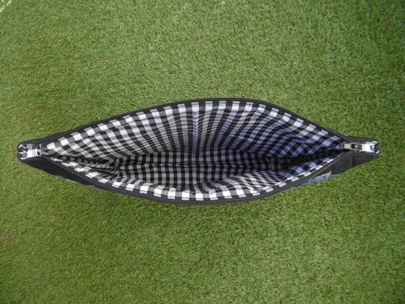 Pu&ograve; includere: Pochette nera con cerniera e fodera a quadretti bianchi e neri. La pochette &egrave; aperta, rivelando il motivo interno. L'esterno sembra essere in tessuto scuro. La pochette &egrave; appoggiata su una superficie erbosa verde.