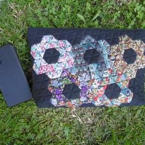Pu&ograve; includere: Pochette trapuntata nera con un design patchwork floreale. Il patchwork presenta forme esagonali con vari motivi floreali nei toni del rosa, viola e blu. Uno smartphone nero &egrave; appoggiato sull'erba accanto alla pochette.