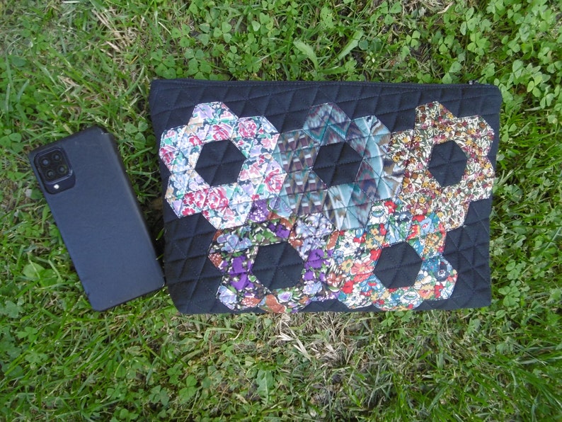 Pu&ograve; includere: Pochette trapuntata nera con un design patchwork floreale. Il patchwork presenta forme esagonali con vari motivi floreali nei toni del rosa, viola e blu. Uno smartphone nero &egrave; appoggiato sull'erba accanto alla pochette.