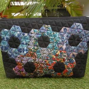 Op de afbeelding: Zwarte gewatteerde etui met ritssluiting en een patchwork ontwerp. Het patchwork heeft zeshoekige bloempatronen in tinten blauw, groen en rood. De etui is rechthoekig en ligt op een groene ondergrond.