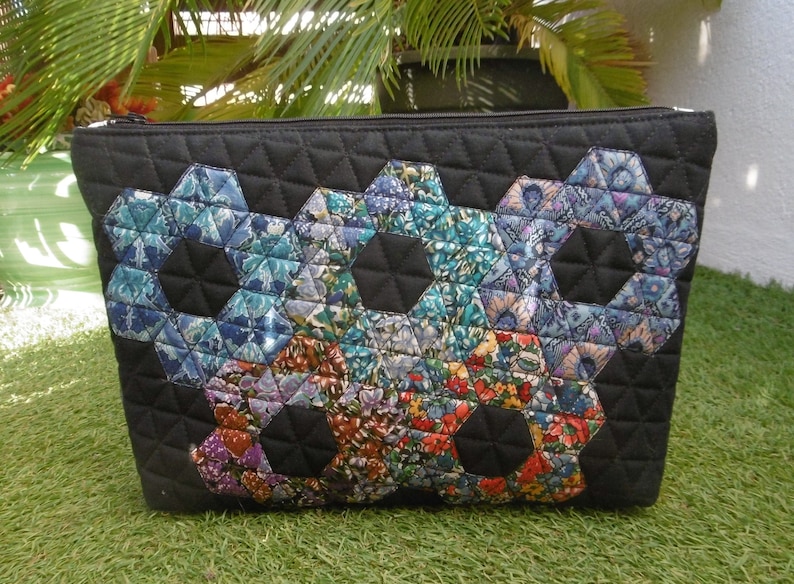 Pu&ograve; includere: Pochette nera trapuntata con cerniera e design patchwork. Il patchwork presenta motivi floreali esagonali nei toni del blu, verde e rosso. La pochette &egrave; rettangolare e poggia su una superficie verde.