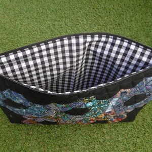 Pu&ograve; includere: Pochette nera con cerniera con un design trapuntato e un colorato motivo patchwork. L'interno &egrave; foderato con un tessuto a quadretti bianco e nero. La pochette &egrave; su uno sfondo verde.