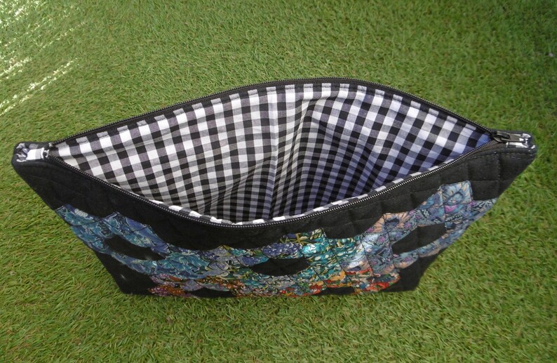 Pu&ograve; includere: Pochette nera con cerniera con un design trapuntato e un colorato motivo patchwork. L'interno &egrave; foderato con un tessuto a quadretti bianco e nero. La pochette &egrave; su uno sfondo verde.