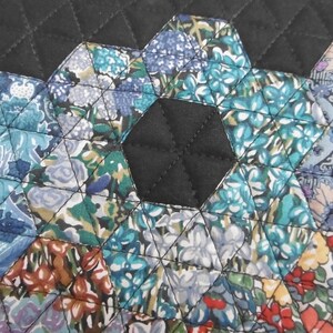 Pu&ograve; includere: Primo piano di un tessuto trapuntato con un motivo geometrico. Il design presenta forme esagonali in varie stampe floreali, tra cui blu, verde acqua e arancione, cucite insieme con filo nero su uno sfondo nero.