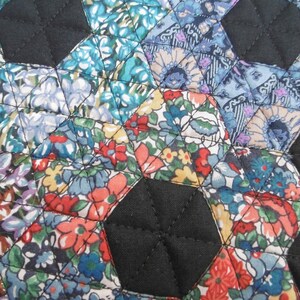 Pu&ograve; includere: Primo piano di un tessuto trapuntato con un motivo floreale nei toni del blu, rosso e giallo. Le forme esagonali sono cucite insieme con filo nero e il tessuto nero riempie gli spazi tra gli esagoni. La trapunta ha un aspetto fatto a mano.