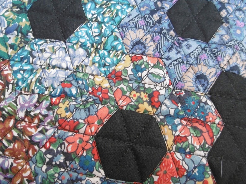 Pu&ograve; includere: Primo piano di un tessuto trapuntato con un motivo floreale nei toni del blu, rosso e giallo. Le forme esagonali sono cucite insieme con filo nero e il tessuto nero riempie gli spazi tra gli esagoni. La trapunta ha un aspetto fatto a mano.