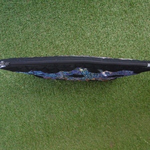 Pu&ograve; includere: Una pochette rettangolare nera con cerniera e un design colorato a mosaico sul davanti. La pochette &egrave; appoggiata su una superficie verde e la cerniera corre lungo il bordo superiore.