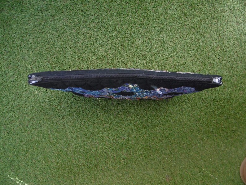 Pu&ograve; includere: Una pochette rettangolare nera con cerniera e un design colorato a mosaico sul davanti. La pochette &egrave; appoggiata su una superficie verde e la cerniera corre lungo il bordo superiore.