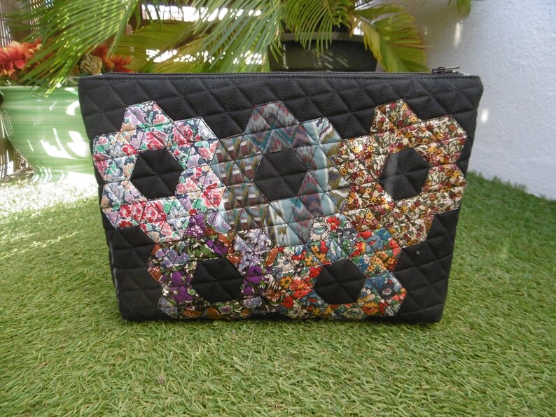 Pu&ograve; includere: Pochette cosmetica trapuntata nera con chiusura a cerniera. La borsa presenta tre grandi disegni esagonali floreali nei toni del rosso, viola e giallo. La borsa &egrave; appoggiata su un letto di erba verde.