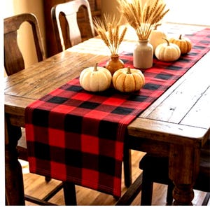 May include: A red and black buffalo plaid table runner featuring a repeating checkered pattern. The runner is decorated with two white pumpkins, one orange pumpkin, and two vases filled with dried wheat stalks, arranged along the center.