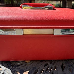 May include: A red vintage travel case with a silver handle and two silver latches. The case has a gold trim around the edge.