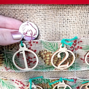 Könnte beinhalten: Ein Adventskalender aus Jute mit Holzschmuck, der von 1 bis 15 nummeriert ist. Der Schmuck ist in Form von Christbaumkugeln und hängt an grünem Garn. Der Kalender ist mit einem grün-weißen Band mit Tannenzapfenmuster verziert.