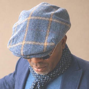 May include: A blue tweed flat cap featuring a prominent yellow and brown windowpane check pattern. The cap is constructed with a textured herringbone weave and has a structured, rounded crown with a short, stiff brim extending from the front.